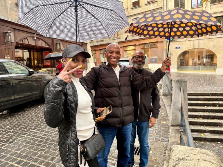 Three people smiling under umbrellas on a rainy cobblestone street in Geneva